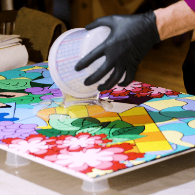 A person in gloves pouring clear liquid onto a red, green, yellow, orange, blue, and purple painting